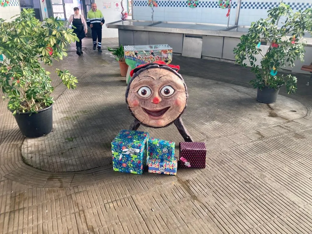 Tió gegant de cara, amb barretina i uns quants regals als peus, en un mercat decorat per Nadal