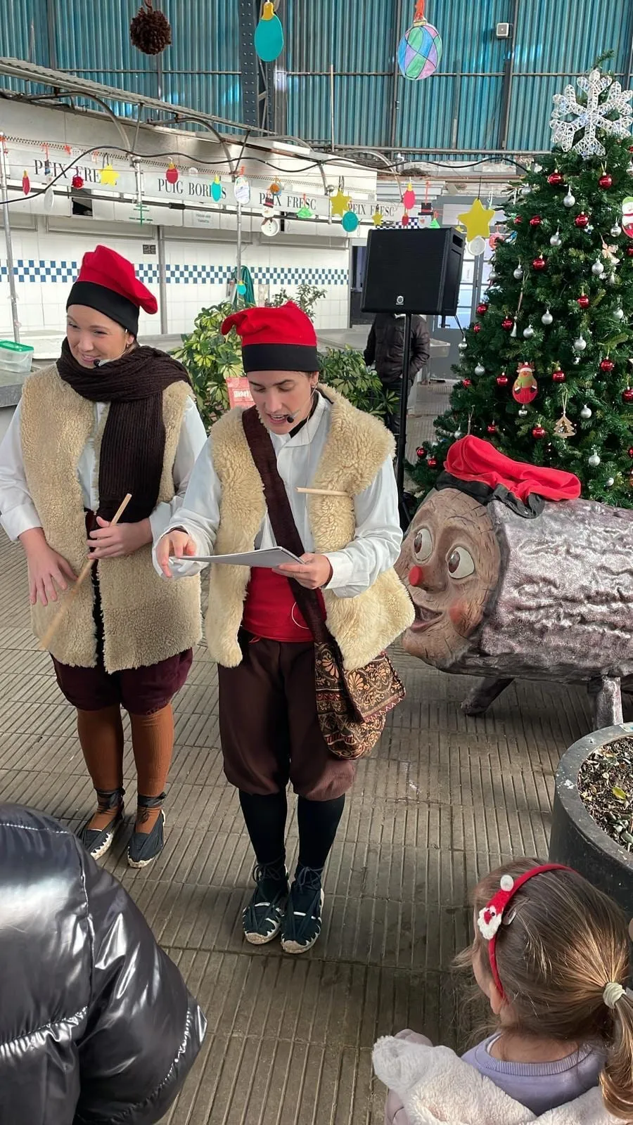 Dues intèrprets amb barretina i armilla de pell, amb bastons, al costat d’un tió gegant en un mercat de Nadal cobert