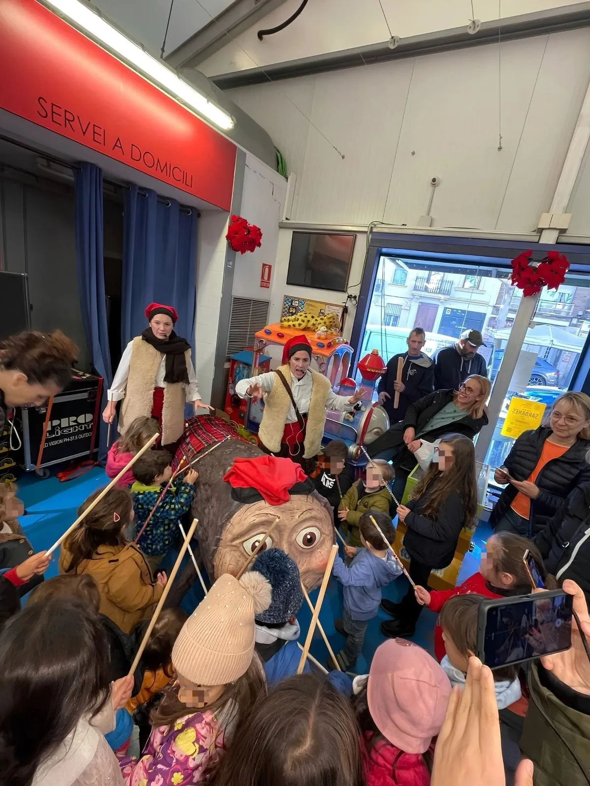 Grup d’infants amb bastons al voltant d’un tió gegant durant l’espectacle, amb les intèrprets al fons