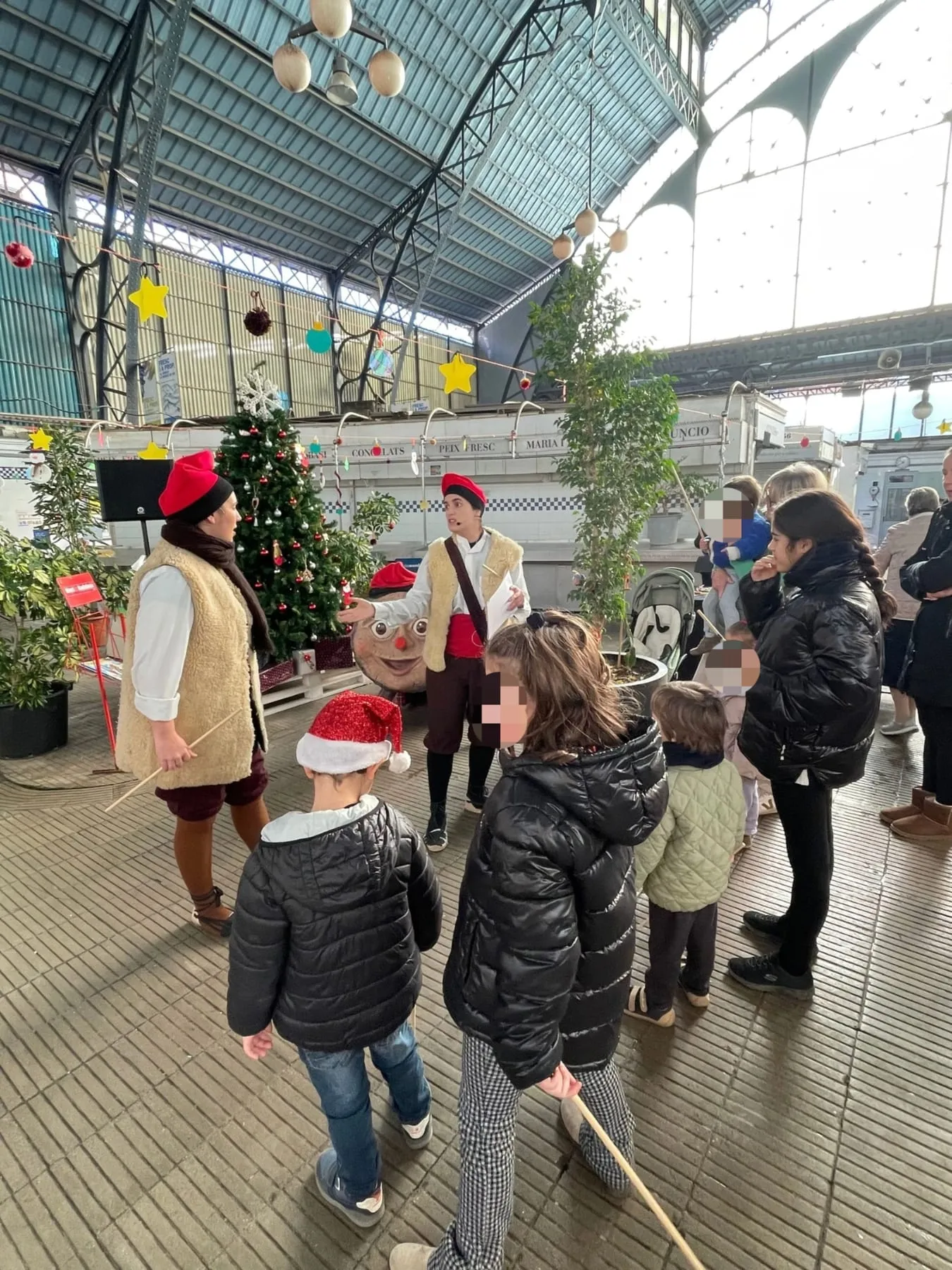 Intèrprets i infants amb bastons en un mercat cobert decorat per Nadal, amb el tió gegant al fons
