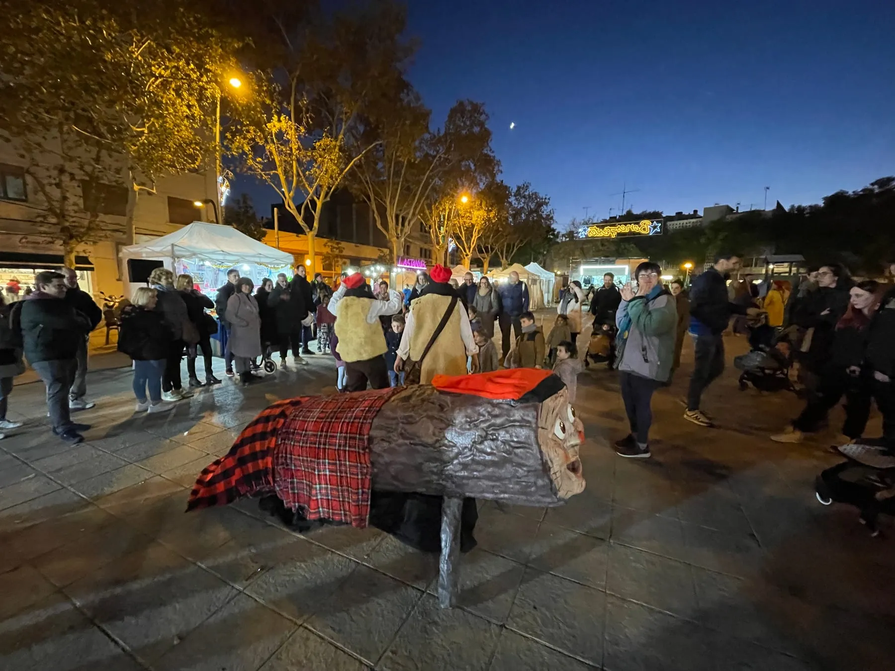 Tió gegant amb manta de quadres a l’aire lliure, de nit, en una fira de Nadal amb famílies