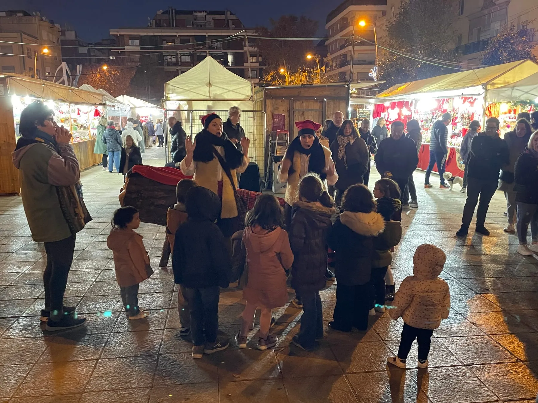 Famílies i infants al voltant del tió gegant en una fira de Nadal nocturna amb parades il·luminades