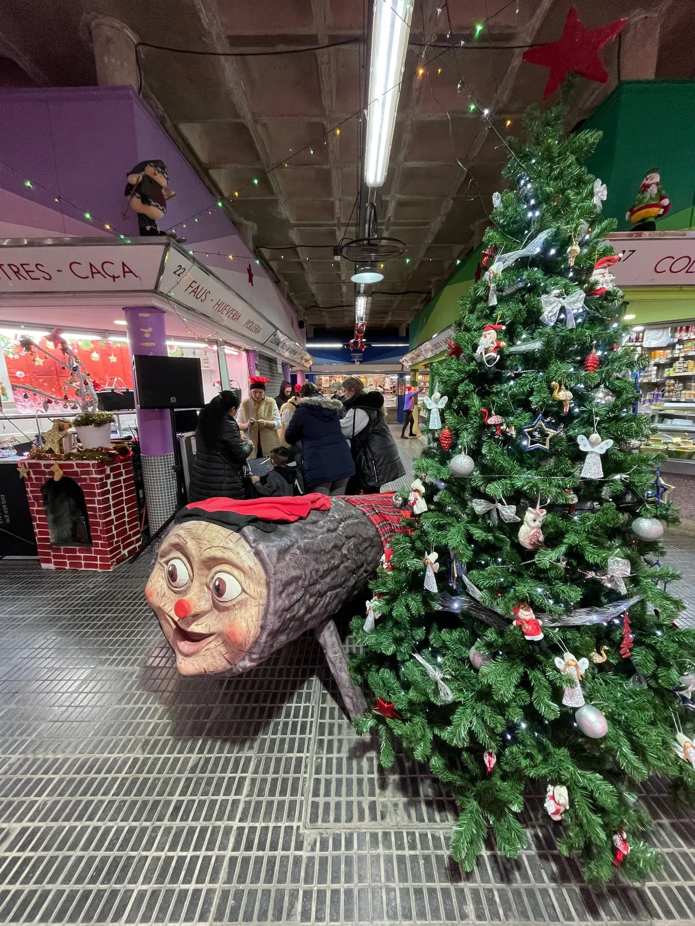 Tió gegant amb barretina al costat d’un avet decorat en un mercat municipal