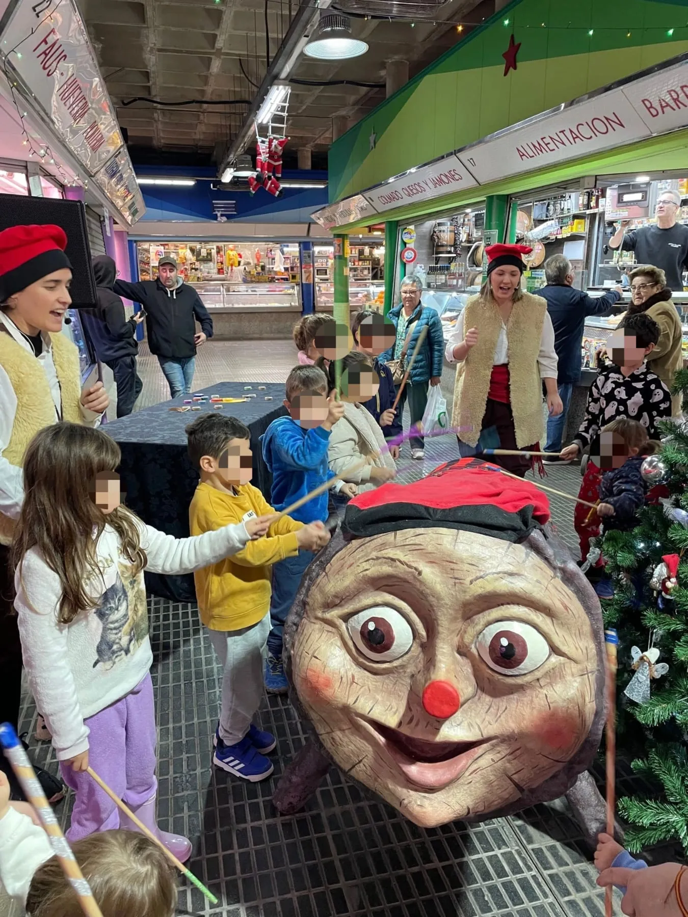 Infants amb bastons fent cagar el tió gegant en un mercat, amb les intèrprets darrere
