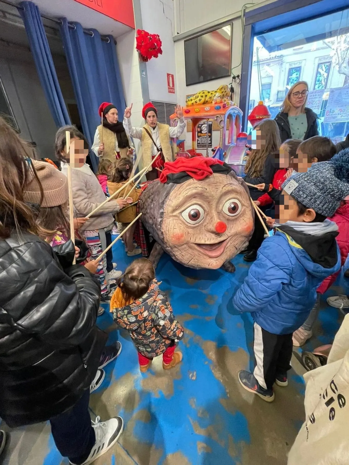Infants amb bastons al voltant d’un tió gegant durant l’espectacle, amb dues intèrprets al fons